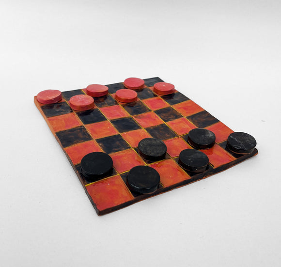 This ceramic sculpture represents a checkers game board with a classic red-and-black checker pattern, featuring red and black playing pieces positioned as if mid-game. The hand-crafted surface and matte finish of the board and pieces highlight the handmade nature of the artwork, adding texture and character to the familiar design.