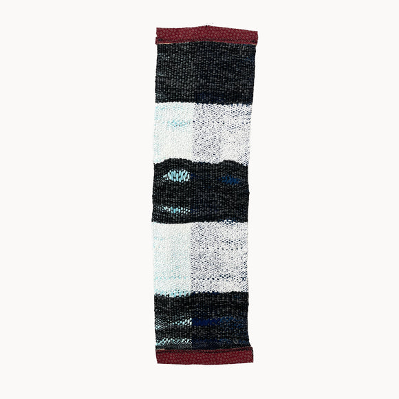 This textile artwork is a rectangular woven piece featuring alternating bands of black, white, and subtle blue threads, with bold red edges at the top and bottom. The textures and patterns within each band create a visual rhythm, drawing attention to the contrast between the monochrome tones and hints of color.