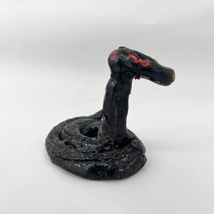 This ceramic sculpture portrays a coiled snake with a raised head, its dark, glossy glaze accentuating the sinuous curves and texture of its body. The piece exudes an ominous presence, with subtle red details near the eyes suggesting a sense of latent aggression or alertness, imbuing the snake with a lifelike and intimidating quality.