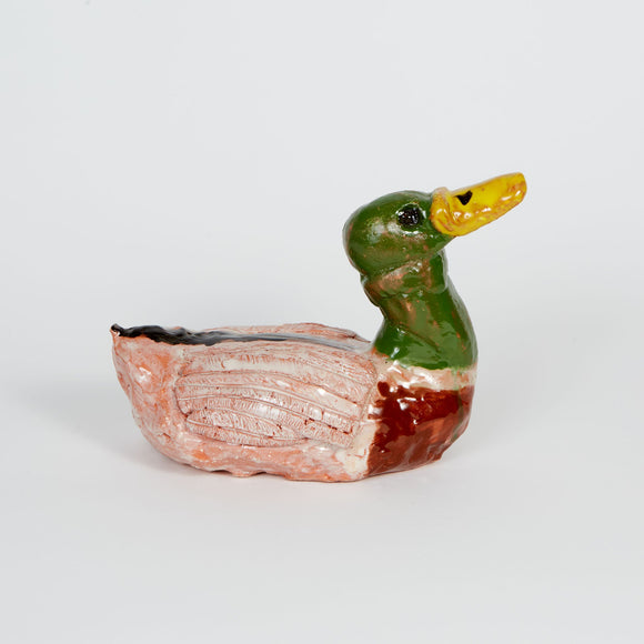 This ceramic sculpture depicts a mallard duck (Anas platyrhynchos) with a characteristic green head, yellow beak, and brown chest, accurately mirroring the male mallard's distinct plumage. The handcrafted texture and color variations highlight the artisanal quality, creating a stylized yet recognizable representation of this common waterfowl.