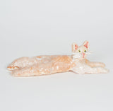This ceramic sculpture depicts a relaxed, elongated cat with a serene expression, crafted in soft tones of cream and light peach. The figure’s stretched body and gentle glaze create a sense of calm and simplicity, emphasizing the cat's peaceful demeanor.