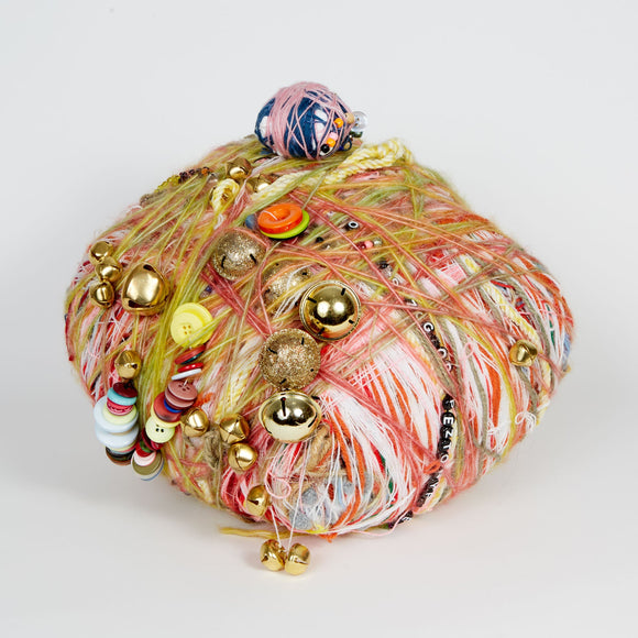 This sculpture is a vibrant assemblage of multicolored yarn, buttons, jingle bells, and beads intricately wrapped into a rounded form. The textured layering of different materials creates a dynamic, playful aesthetic, invoking themes of craft, nostalgia, and ornamentation.