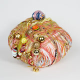 This sculpture is a vibrant assemblage of multicolored yarn, buttons, jingle bells, and beads intricately wrapped into a rounded form. The textured layering of different materials creates a dynamic, playful aesthetic, invoking themes of craft, nostalgia, and ornamentation.