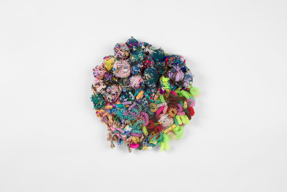 This round, mixed media work consists of several spherical weavings of yarn, tightly rolled strips of fabric, and spiral hair ties in a variety of colors including pastel pink, warm purple, cool teal, lime green, and hot pink.